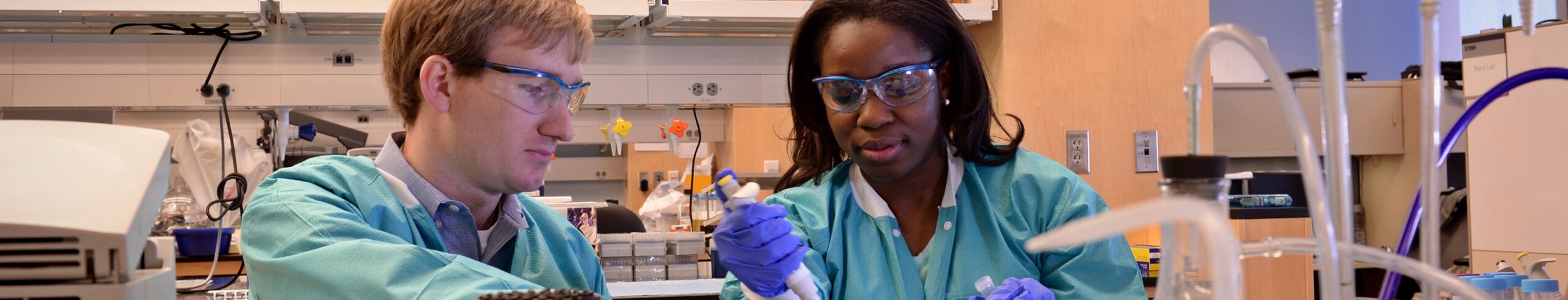 Photo of two researchers in the lab wearing PPE gear and pipetting.