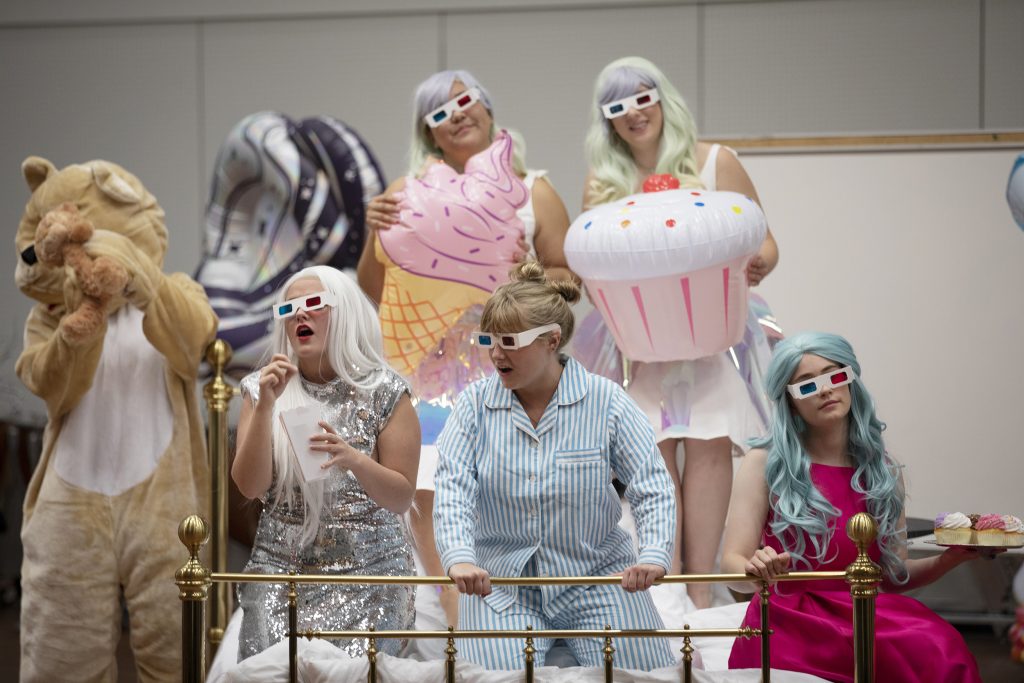 Five women dressed in dresses, sequins and pajamas sit on a bed in Kenan Music Building