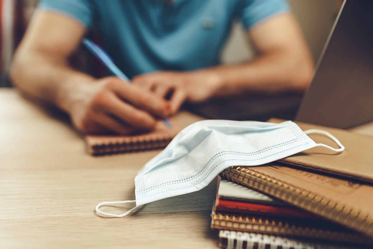 Photo: Male student studies at home during quarantine. Close up of a table with laptop, notebooks and medical mask.