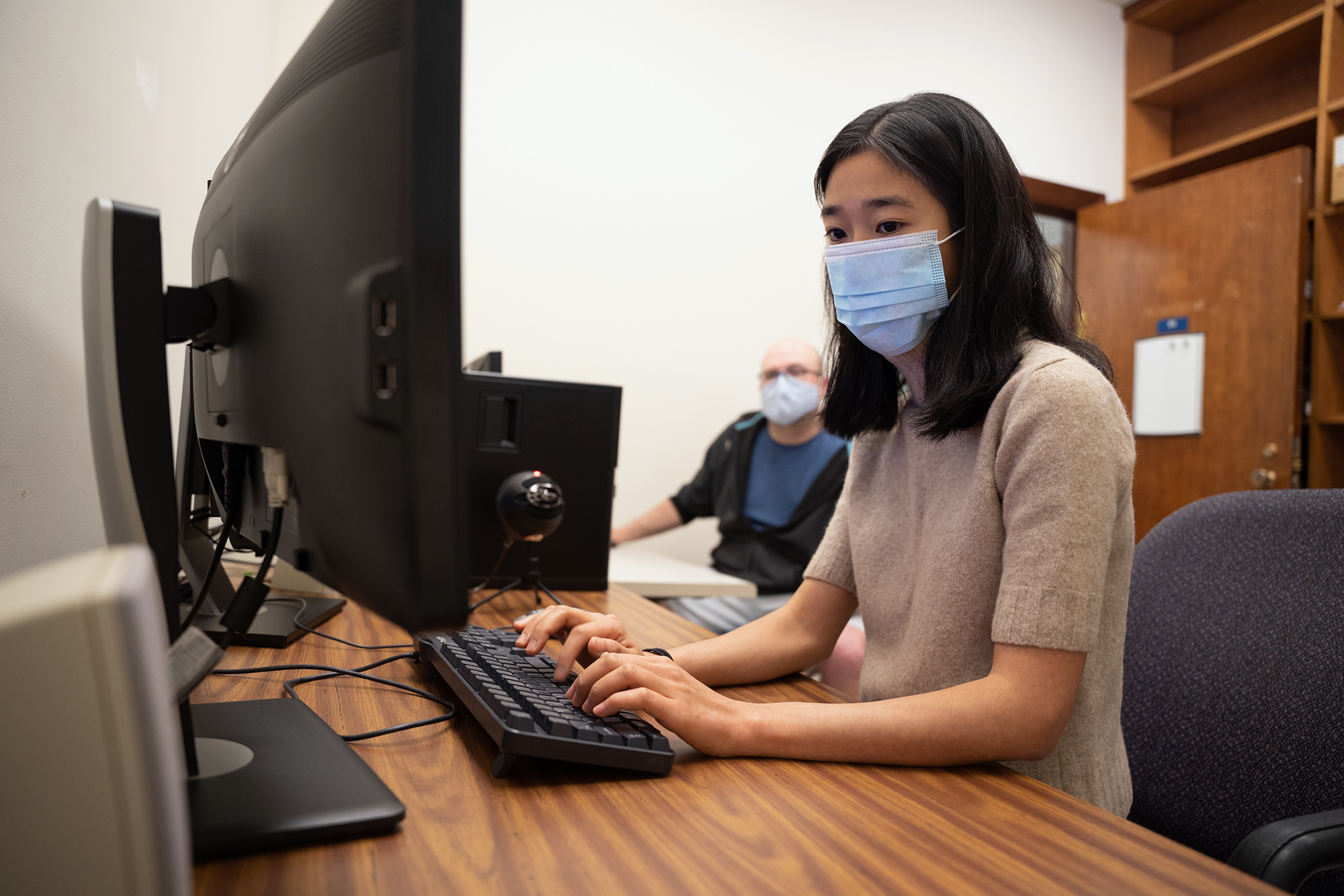 A woman types at a computer while a man seated behind her looks on.