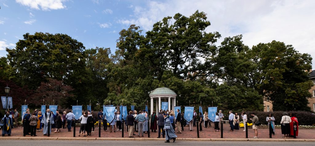 Photo of people in graduation robs, holding Carolina blue banners lined up near the Old Well.