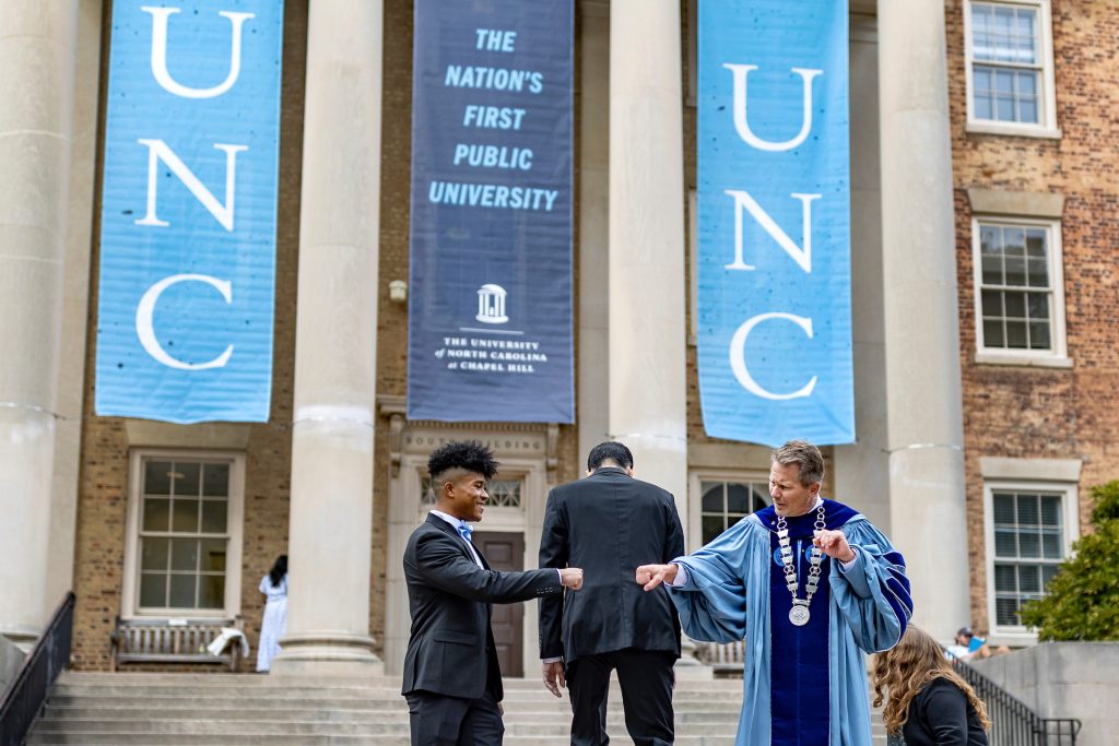 Kevin Guskiewicz, wearing Carolina blue graduation robes, fist bumps Christian Chung outside a building that has a banner that reads "The Nation's First Public University."