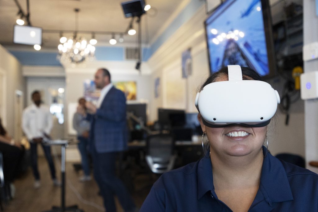 A person wears a VR headset while they're smiling, presenters are talking in the background.