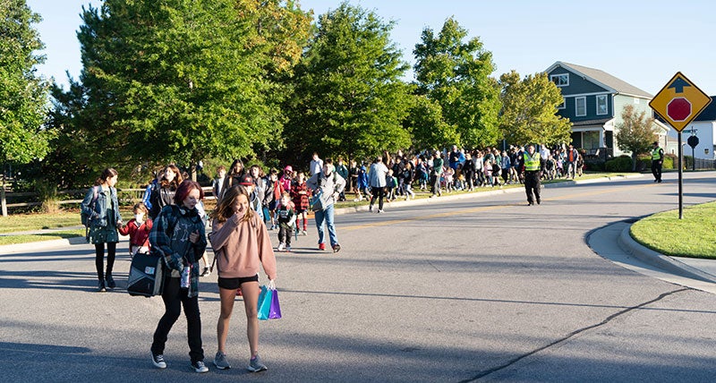 A long line of students with some parents walking to school on a sidewalk and crossing the road.