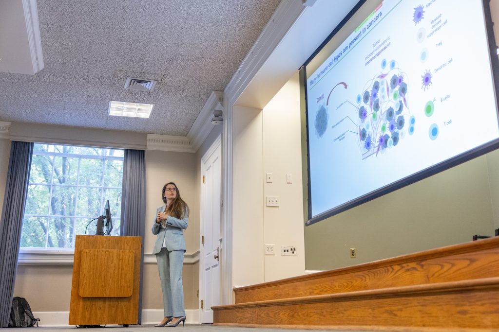Yuliya Pylayeva-Gupta stands at a podium at the front of the room, she looks to the side at a slide that reads "Many Immune Cell Types are Present in Cancers."