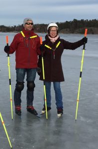 Two people in cold weather gear with ice skates on, standing on a frozen lake.