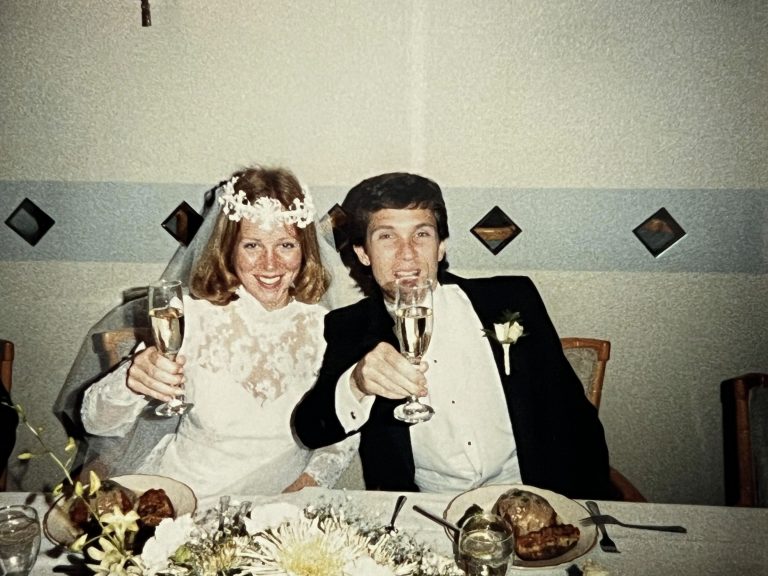Photo so Bulik and Sullivan at their wedding. They're smiling at the camera, holding wine glasses in a cheers like fashion.