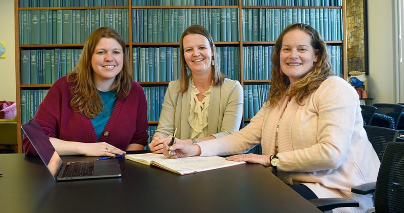 Ashley Melzer, Courtney Rivard, and Jordynn Jack sitting at a table in front of a bookcase filled with green books