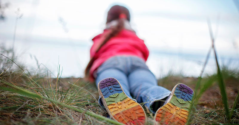 A young person laying in the grass with the bottom of their shoes shown in full view. The shoes have a rainbow painted on the bottoms.