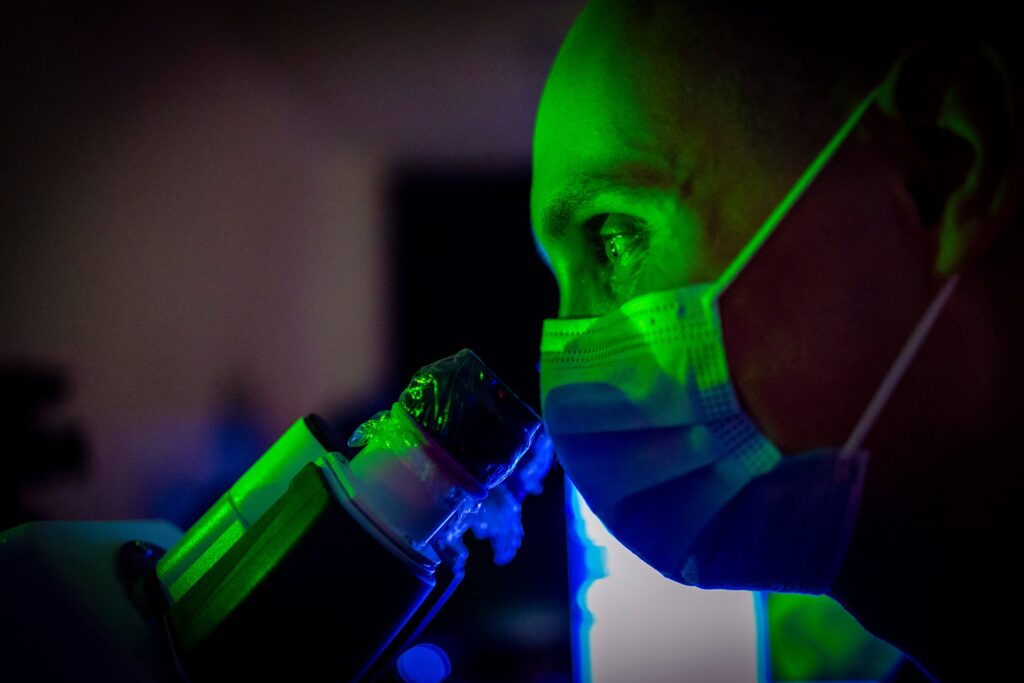 A person wearing a medical facemask peers into a telescope under a green light.