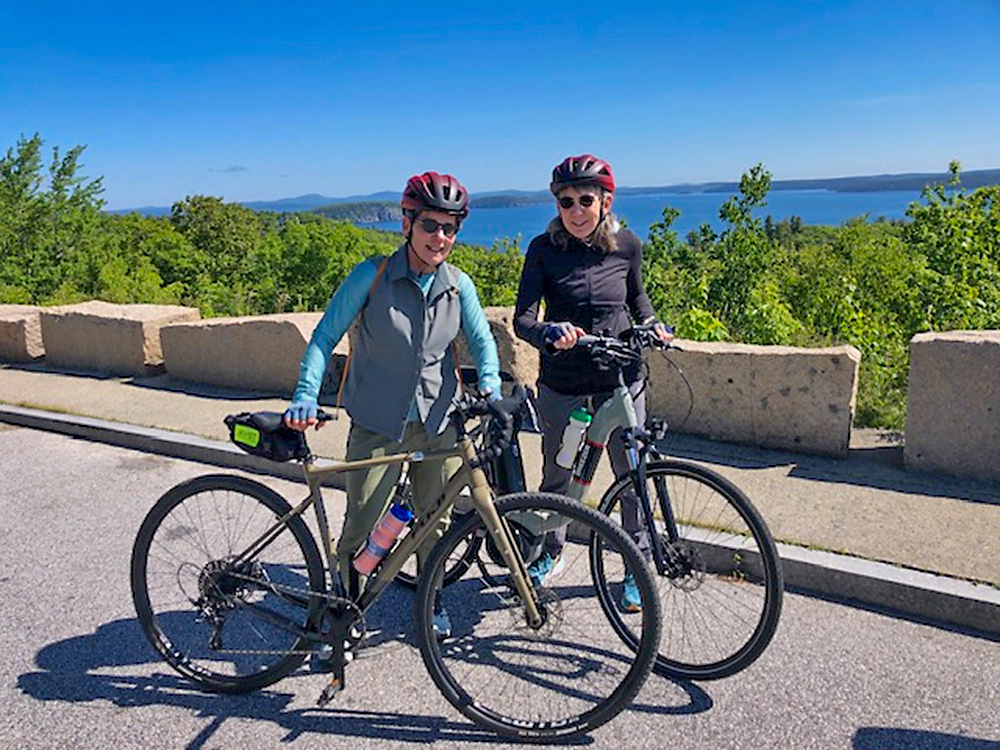 Victoria Rovine and Florence Babb pose for a photo with their bikes in front of a lake surrounded by trees.