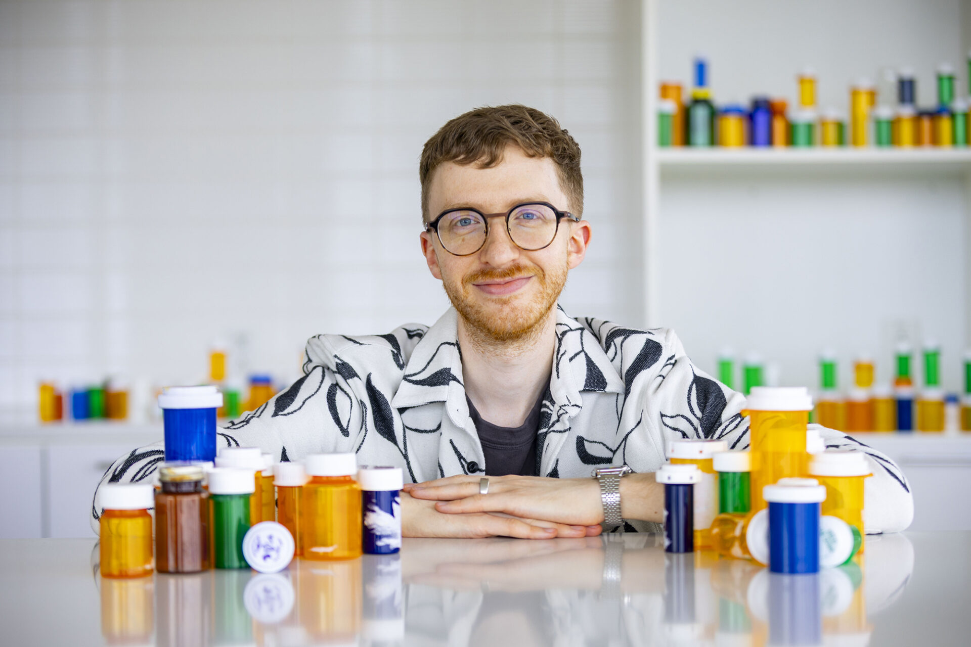 a young man leaning against a white table covered in colorful prescription bottles
