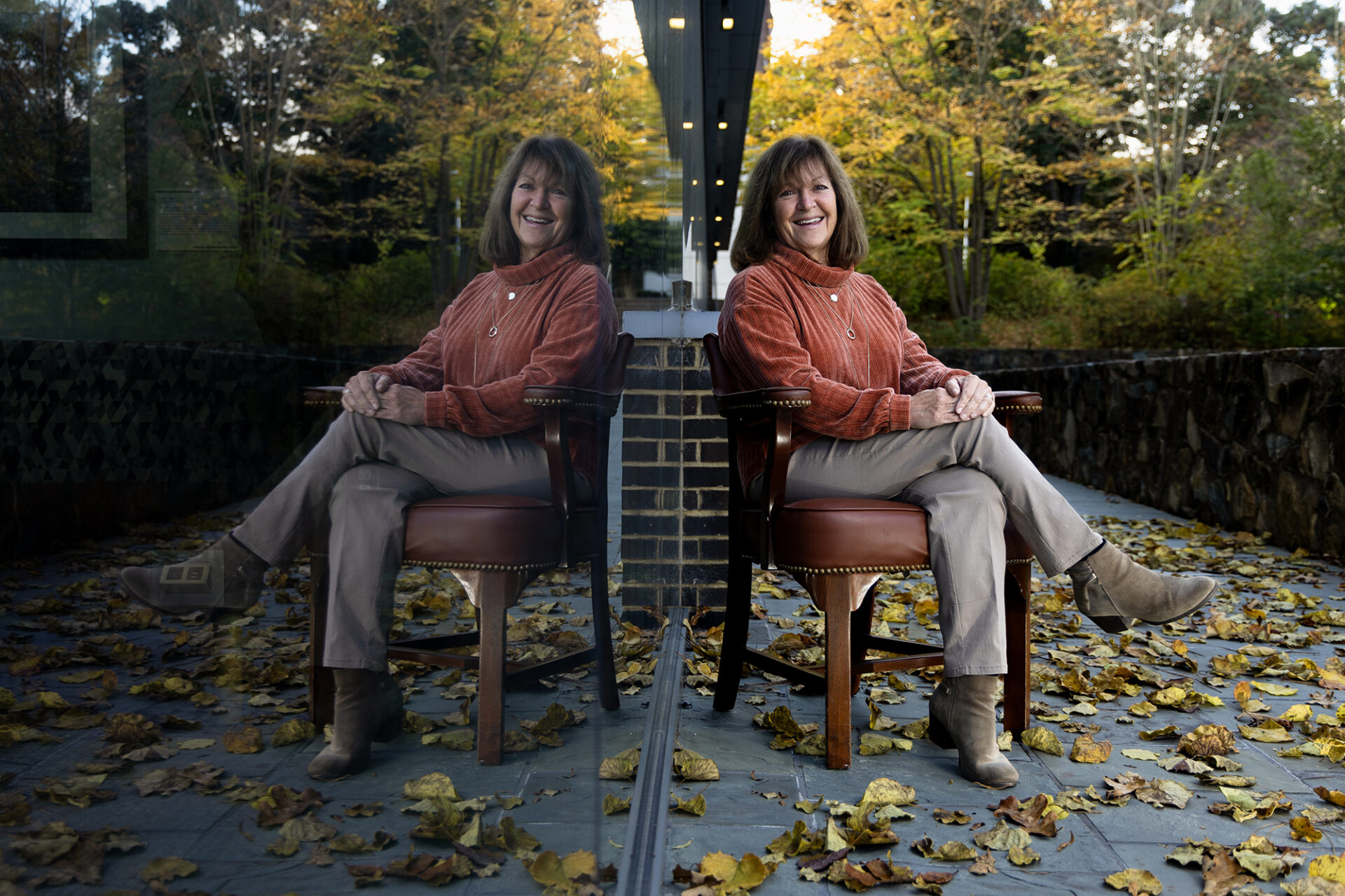 a woman sits in a chair surrounded by leaves, her reflection in a nearby window