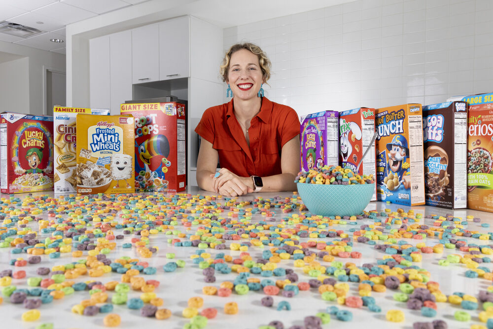 A woman in a red shirt smiles between several boxes of breakfast cereal. The counter in front of her is also covered in loose cereal.