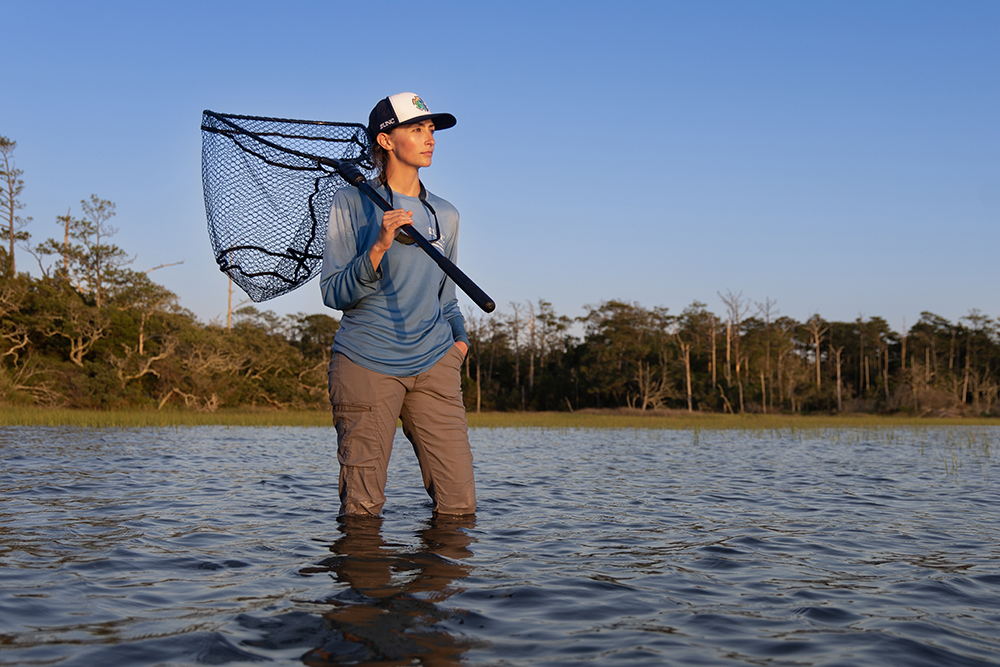 young woman holding a fishing net over her shoulder looks out at the water