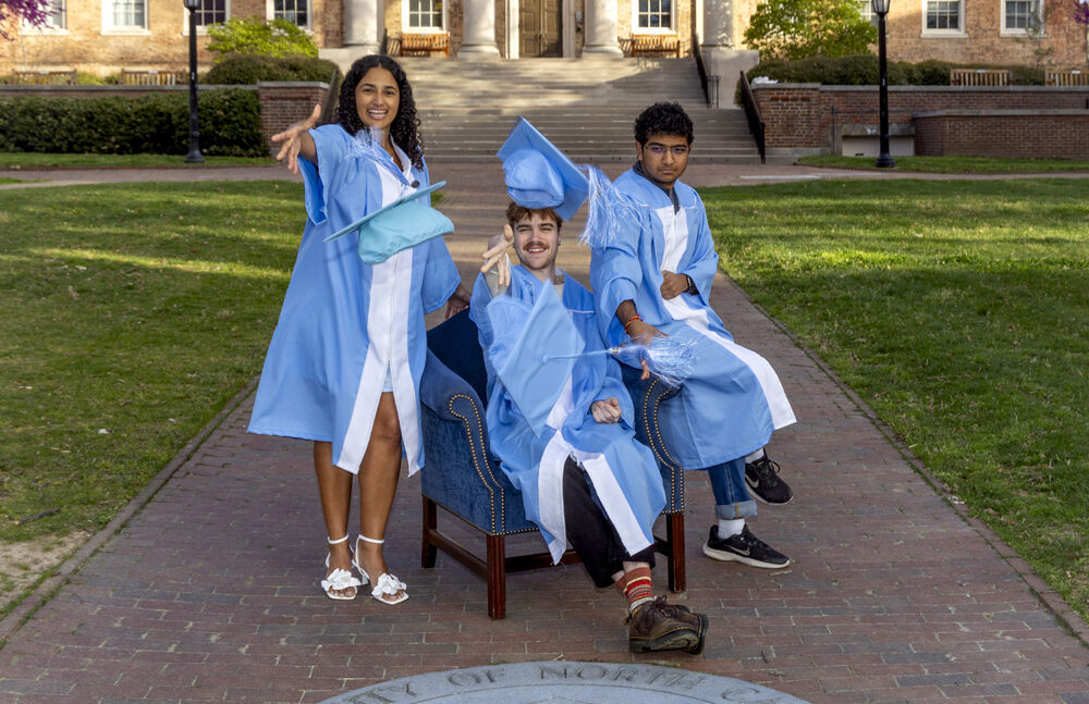 Photo of Kiara Rodriguez, Nico Gleason, and Karthik Ramakrishnan in front of Smith Building in their cap and gowns, throwing their caps at the camera.