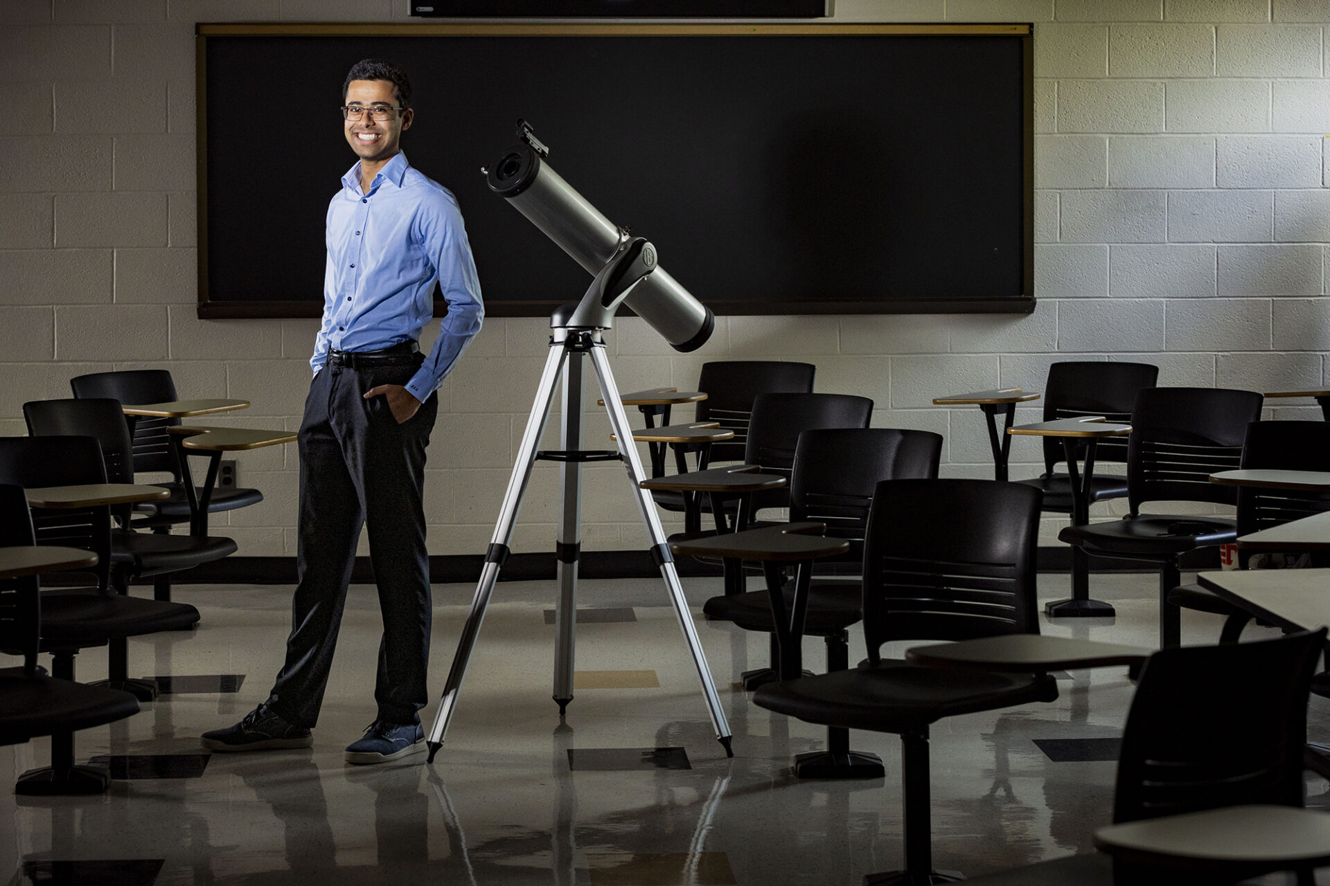 a young man stands next to a telescope in a classroom