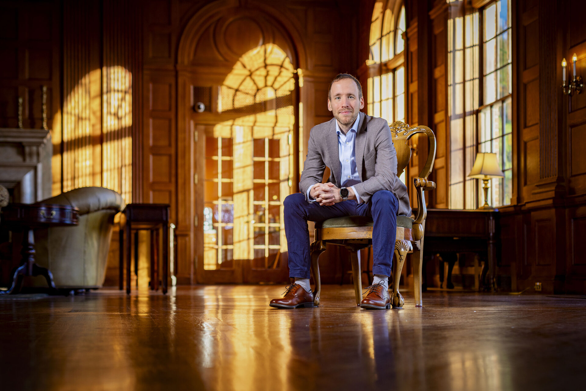 a man sits in a wingback chair in a wood-paneled room with big windows