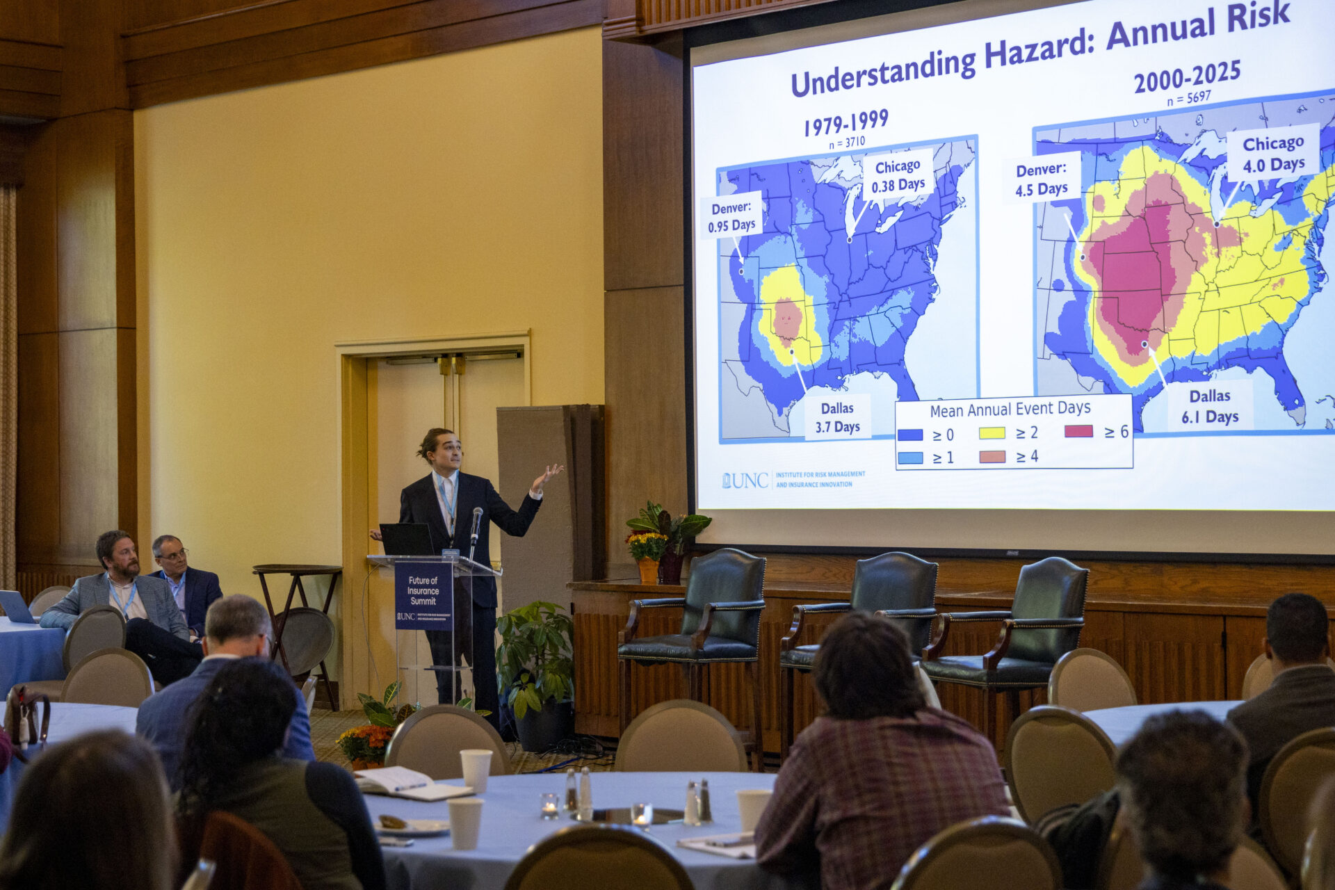 A man in a large conference room gesturing to images on a projection screen.