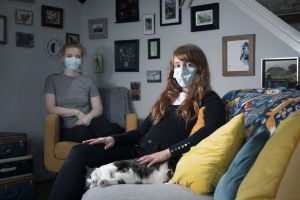 two young women wear masks as they sit in their living room