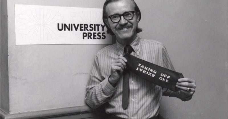Photo: A smiling man in 1970s-era attire holds up his necktie, which has been stenciled with the words 'Taking Off.' He stands in front of a sign that reads 'University Press.'