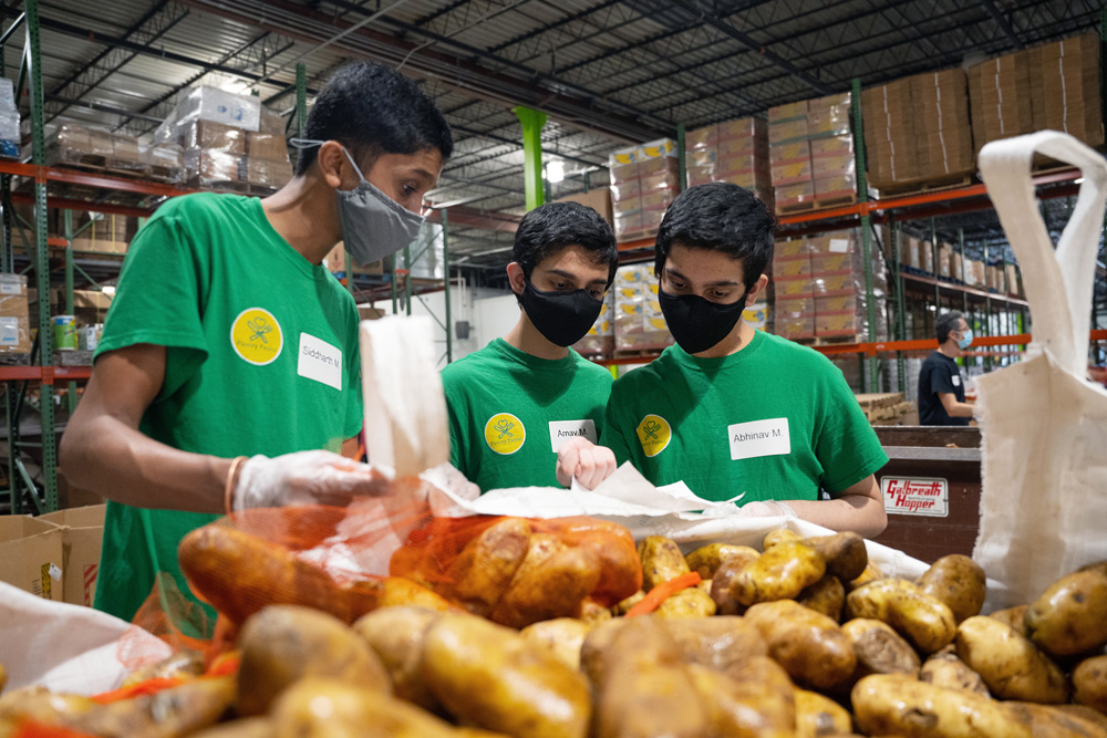 Three students wearing masks and green “Pantry Patrol” t-shirts examine paperwork behind a bin of potatoes in a warehouse.