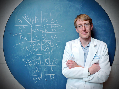 Photo: A man in a white lab coat stands with his arms crossed in front of a circular blue chalkboard covered with genetics diagrams.