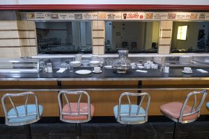 A mid-century diner counter with alternating pink and baby-blue stools. Several stacks of white dishes are visible. A row of placards above the counter advertise items such as “Cherry Pie, 15 cents” and “Roast Turkey Dinner, 65 cents.”
