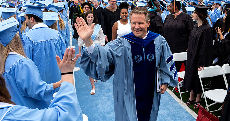A man in an academic robe at a college graduation high-fiving a student in a graduation gown.